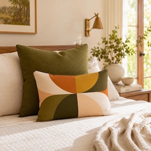 May include: A square olive green throw pillow and a rectangular lumbar pillow featuring a geometric pattern of cream, orange, and mustard yellow semi-circles. The pillows are arranged on a bed with a white textured quilt and a cream-colored pillow.