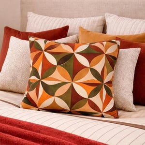 May include: A collection of decorative throw pillows in various shapes and colors. The central pillow features a geometric pattern in orange, brown, green, and cream. Other pillows are solid in shades of rust, cream, and gold, creating a cozy, inviting bedroom setting.