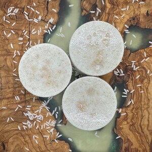 Può includere: Tre saponette rotonde, bianche, con una texture granulosa, su una superficie di legno con riso bianco sparso intorno.
