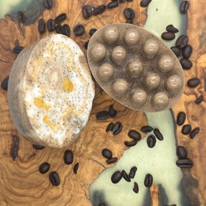 Può includere: Due saponette profumate al caffè su una superficie di legno con chicchi di caffè sparsi intorno. Una saponetta è rotonda, marrone, con un motivo a spirale e brillantini dorati. L'altra saponetta è marrone, con una superficie testurizzata e una griglia di cerchi in rilievo.