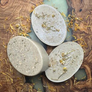 May include: Three oval-shaped bars of soap with a light brown color and flecks of white. The soap bars have dried flowers and herbs embedded in them. The soap bars are sitting on a wooden surface with yellow flower petals scattered around them.