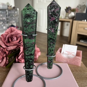 Può includere: Due punte di pietre preziose verdi e nere con accenti rosa su supporti in metallo. I supporti sono su una superficie rosa.