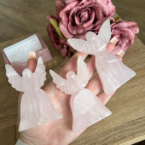 Puede incluir: Tres figuritas de &aacute;ngel de cristal rosa con detalles tallados. Los &aacute;ngeles est&aacute;n dispuestos en una mano con un reloj blanco.