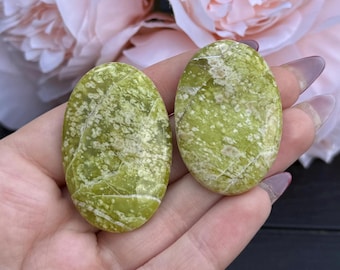 Puede incluir: Dos piedras ovaladas verdes con vetas blancas. Las piedras tienen un aspecto moteado y se sostienen en una mano. Flores rosas suaves en el fondo.