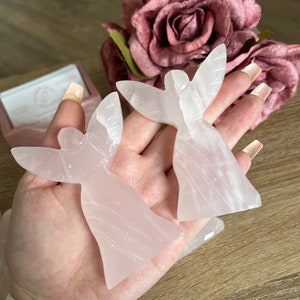 Puede incluir: Dos figuras de &aacute;ngeles rosas talladas en cuarzo rosa. Las figuras tienen los brazos levantados en posici&oacute;n de oraci&oacute;n.