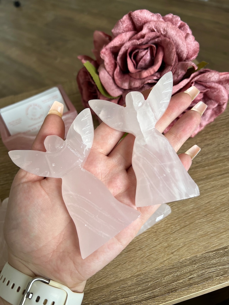 Puede incluir: Dos figuras de &aacute;ngeles rosas talladas en cuarzo rosa. Las figuras tienen los brazos levantados en posici&oacute;n de oraci&oacute;n.