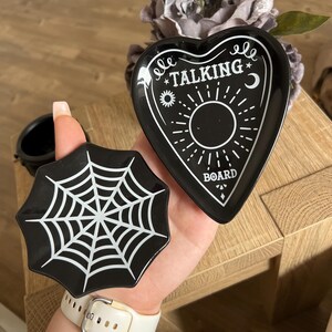 Op de afbeelding: Twee zwarte keramische onderzetters. Eén is hartvormig met een wit "Ouija Talking Board" ontwerp. De andere is rond met een wit spinnenwebontwerp.