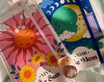 May include: Two rectangular journals featuring tarot card designs. The left journal displays a pink background with a sun face, sunflowers, and the text XIX The Sun. The right journal shows a blue background with a moon face, clouds, and XVIII The Moon.