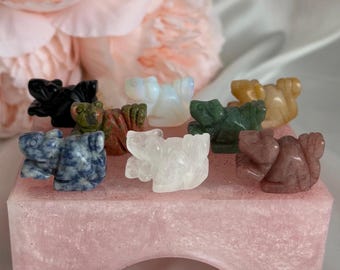 Puede incluir: Una colección de pequeñas figuras de ranas talladas en varios colores, incluyendo negro, verde y blanco, exhibidas sobre una base de resina rosa brillante. El fondo presenta una flor rosa suave.
