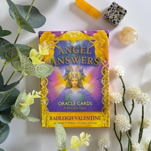 Pode incluir: Um baralho de cartas de oráculo "ANGEL ANSWERS" com uma ilustração de mulher na frente. O baralho é cercado por elementos decorativos, incluindo um cristal amarelo, uma pedra salpicada e uma pedra transparente. O baralho é um baralho de 44 cartas de Radleigh Valentine.