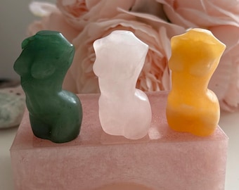 May include: Three carved gemstone figurines in green, pink, and yellow, depicting the female form. They are displayed on a pink resin base. The background features soft pink flowers, creating a serene and artistic composition.