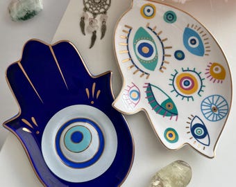 May include: Two decorative ceramic trays. One is a blue Hamsa hand shape with a central eye design, and the other is a white, eye-shaped tray with colorful eye patterns. Both have gold trim.