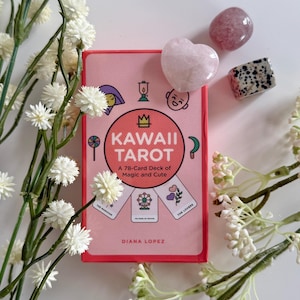 May include: A pink Kawaii Tarot card deck with the text "KAWAII TAROT" and "A 78-Card Deck of Magic and Cute." The deck is surrounded by white flowers and three crystals: a heart-shaped pink crystal, a purple crystal, and a black and white speckled crystal.