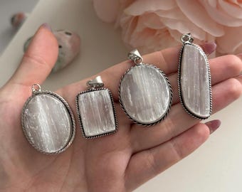 May include: Five selenite crystal pendants in various shapes: oval, rectangular, and circular. Each pendant has a silver-colored metal frame. The crystals are white and translucent. The pendants are displayed on a hand, with a soft pink flower in the background.