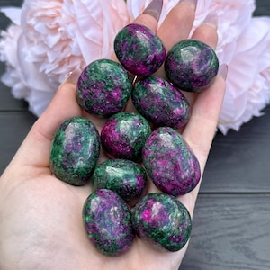 May include: A selection of polished ruby in zoisite stones, each showing a blend of deep green and vivid magenta colours. The stones are smooth and oval, with a natural, speckled look. The stones are held in a hand, with a soft pink flower in the background.