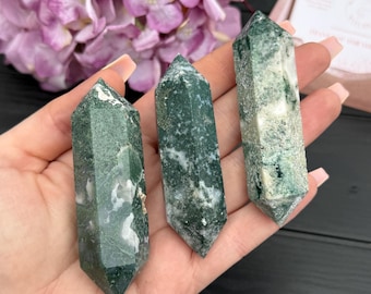 May include: Three green moss agate crystal points with white inclusions are held in a hand. The crystals are polished and have a hexagonal shape, with a pointed top. The background includes pink flowers and a wooden surface.