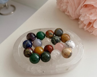 Puede incluir: Una colección de pequeñas esferas de piedras preciosas pulidas y multicolores dispuestas sobre un plato de cristal transparente de forma ovalada. Las esferas presentan varios colores, incluidos verde, rojo, azul, marrón, blanco y rosa.
