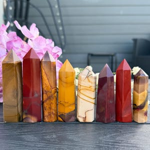 May include: Eight polished gemstone obelisks in a row, featuring colours like brown, red, yellow, and cream. Each obelisk has a pointed top and a rectangular base. The gemstones are displayed against a blurred floral backdrop.