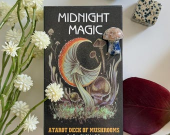 May include: A black tarot card deck box with the words "MIDNIGHT MAGIC" and "ATAROT DECK OF MUSHROOMS" printed on it. The box features a mushroom illustration with orange, blue, and brown colors. Two small stones and a mushroom figurine are also visible.