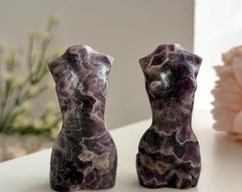 May include: Two polished amethyst stone carvings shaped as human torsos. The sculptures feature deep purple and white crystalline patterns with a smooth, glossy finish. Each piece displays natural mineral variations in dark violet and opaque white hues.