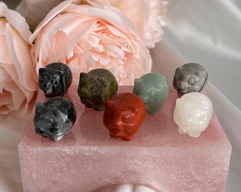 May include: A set of eight small carved animal figurines made of different colored stones, displayed on a pink, glittery base. The figurines are arranged in a semi-circle and include a black and white cat, a green pig, a red owl, a green frog, a white sheep, a gray cat, a green frog, and a white sheep.