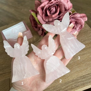 Puede incluir: Tres figuras de &aacute;ngeles rosas talladas en cuarzo rosa. Los &aacute;ngeles est&aacute;n dispuestos en fila sobre una mano, con los n&uacute;meros 1, 2 y 3 escritos debajo de cada uno.