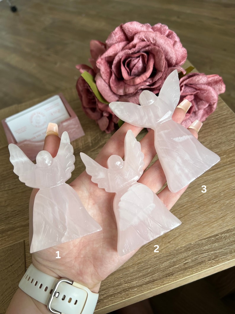 Puede incluir: Tres figuras de &aacute;ngeles rosas talladas en cuarzo rosa. Los &aacute;ngeles est&aacute;n dispuestos en fila sobre una mano, con los n&uacute;meros 1, 2 y 3 escritos debajo de cada uno.