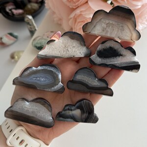 May include: Seven cloud-shaped agate stones in shades of grey, white, and brown. Each stone displays unique patterns and textures. The stones are held in a hand, with a white watch strap visible. The background includes pink flowers.