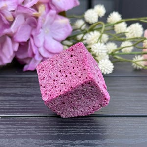Może przedstawiać: Kwadratowy, porowaty przedmiot w kolorze magenta. Obiekt ma teksturowaną powierzchnię z licznymi małymi otworami. Tło obejmuje różowe i białe kwiaty na ciemnej drewnianej powierzchni.