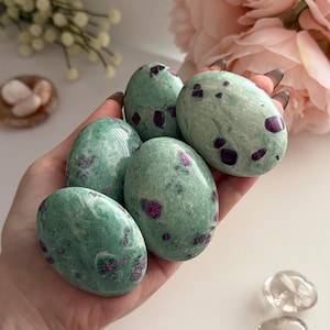 May include: Five polished, oval-shaped stones in shades of green with red spots are held in a hand. The stones have a smooth, natural appearance. The background features soft pink flowers and small white flowers.