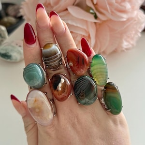 Op de afbeelding: Een hand met negen edelsteenringen. De ringen hebben ovaalvormige stenen in verschillende kleuren, waaronder groen, bruin, oranje en wit, gezet in zilverkleurig metaal. Op de achtergrond een roze bloem.