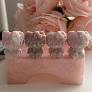 Puede incluir: Cuatro pequeñas figuras de Hello Kitty rosas están dispuestas sobre una base rectangular rosa a juego. Las figuras tienen un aspecto texturizado, similar a la piedra. Suaves flores rosas en el fondo.