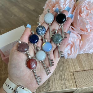 Puede incluir: Un conjunto de ocho colgantes de llave de plata con cabujones de piedras preciosas de diferentes colores. Las piedras preciosas incluyen blanco, negro, azul, verde, rosa y rojo.