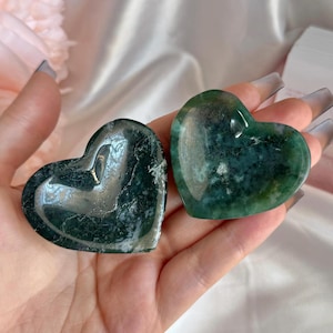 May include: Two heart-shaped stones with green and white patterns are held in a hand. The stones have a polished, smooth surface. The background is a soft, white fabric.