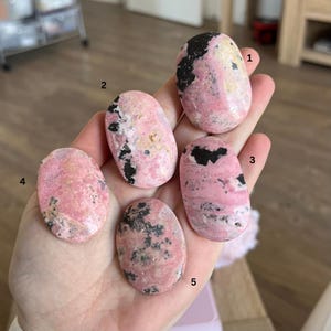 Pu&ograve; includere: Cinque pietre rotonde rosa e nere, note anche come rodocrosite, sono disposte in una mano. Le pietre hanno una forma ovale e una finitura liscia e lucida.
