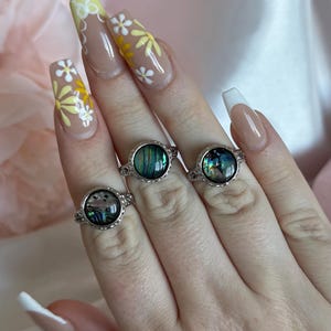 May include: Three silver rings with iridescent, oval-shaped stones are shown. The rings have a decorative, filigree-style setting. The background is a soft pink and white, with a focus on the rings and the hand wearing them.