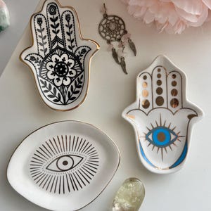 Puede incluir: Colección de cuencos de cerámica blanca con ribete dorado. Los cuencos tienen forma de mano, un óvalo con un diseño de ojo y una mano con un diseño de ojo. También hay un atrapasueños y un cristal.