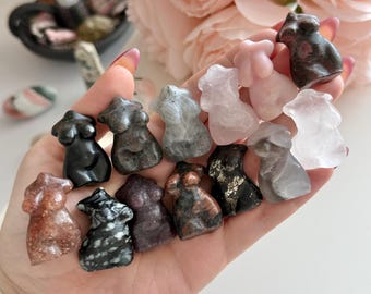May include: A selection of small, carved crystal figurines shaped like a female torso. The crystals are in various colours, such as black, grey, pink, and brown. The figurines are held in a hand, with pink flowers in the background.