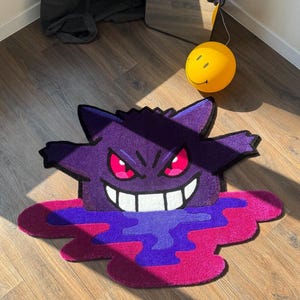 Puede incluir: Una alfombra de mechones con forma personalizada que presenta al personaje morado Gengar con orejas puntiagudas, ojos rojos brillantes y una amplia sonrisa blanca. El personaje parece derretirse en un charco vibrante de tonos morado y magenta.