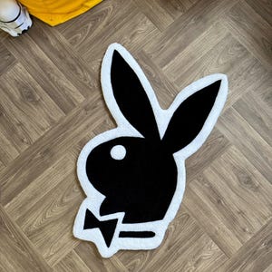Puede incluir: Una alfombra blanca con un logotipo de conejito Playboy negro en el centro. El conejito lleva una pajarita.