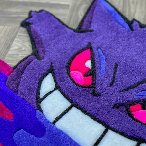 Puede incluir: Una alfombra tufting con la forma del personaje Gengar, que presenta un cuerpo de color morado intenso con una amplia sonrisa de dientes blancos, ojos de color rosa brillante y detalles en azul oscuro. Tiene un pelo grueso y texturizado.