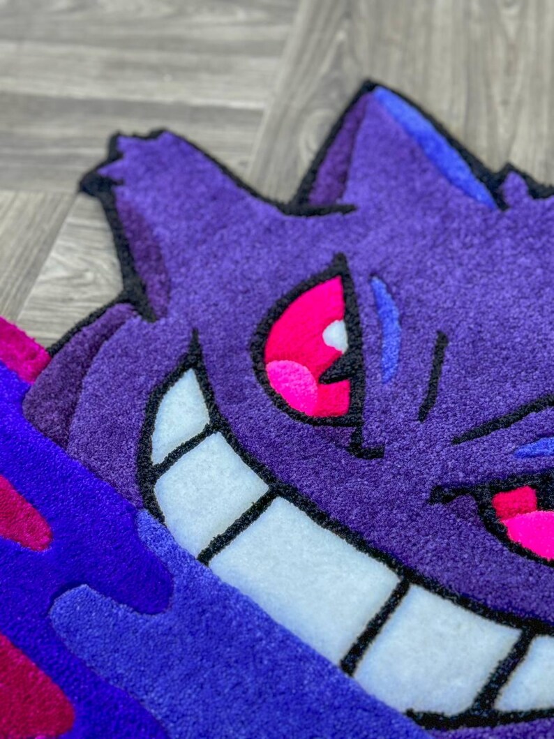 Puede incluir: Una alfombra tufting con la forma del personaje Gengar, que presenta un cuerpo de color morado intenso con una amplia sonrisa de dientes blancos, ojos de color rosa brillante y detalles en azul oscuro. Tiene un pelo grueso y texturizado.