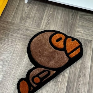 May include: A brown and black rug with a cartoon monkey design. The monkey's face is orange and the rug is made of a soft, plush material.