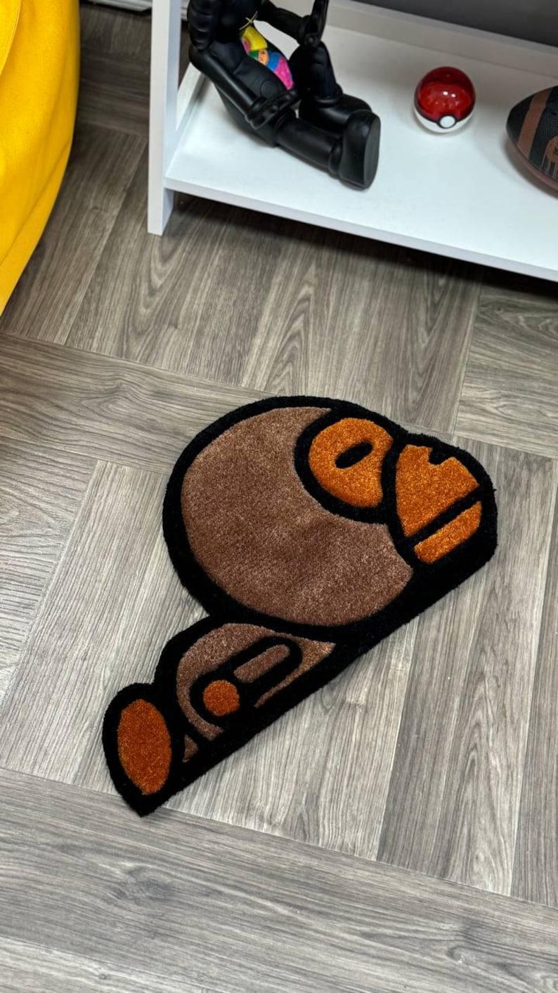 May include: A brown and black rug with a cartoon monkey design. The monkey's face is orange and the rug is made of a soft, plush material.