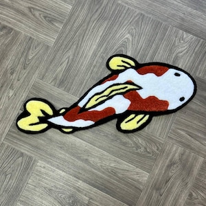 May include: A red, white, and yellow plush rug shaped like a koi fish. The rug has a black outline and is made of soft, fuzzy material.