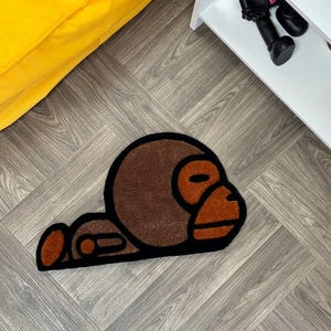 May include: A brown and black rug with a cartoon monkey design. The monkey is lying on its side with its head resting on its arm.