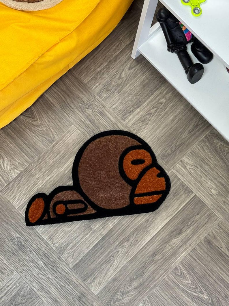 May include: A brown and black rug with a cartoon monkey design. The monkey is lying on its side with its head resting on its arm.