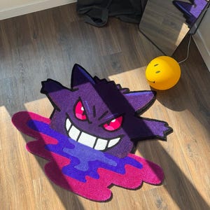 Puede incluir: Una alfombra de tufting con la forma del personaje Gengar, que presenta un cuerpo de color morado intenso, ojos de color rosa brillante y una amplia sonrisa blanca. El personaje parece emerger de un charco vibrante y arremolinado de l&iacute;quido morado y magenta.