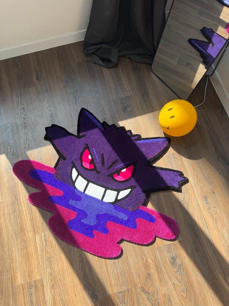 Puede incluir: Una alfombra de tufting con la forma del personaje Gengar, que presenta un cuerpo de color morado intenso, ojos de color rosa brillante y una amplia sonrisa blanca. El personaje parece emerger de un charco vibrante y arremolinado de l&iacute;quido morado y magenta.
