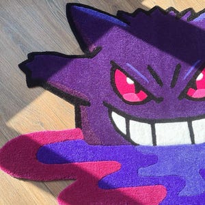 Puede incluir: Una alfombra tufting con la forma del personaje Gengar, que presenta un cuerpo de color morado intenso con un contorno negro, ojos de color rosa brillante y una amplia sonrisa blanca. La base muestra un dise&ntilde;o abstracto de charco en tonos magenta y violeta.